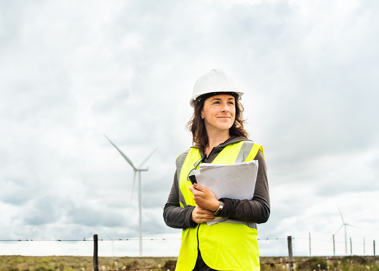 A woman in a hard hat and hi-visibility vest stands in front of wind turbines