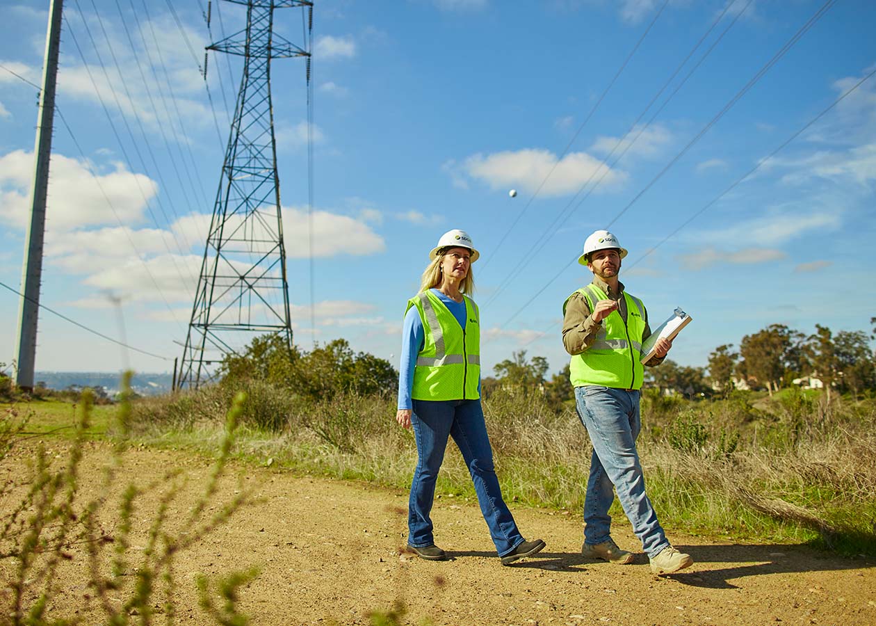 Two Sempra workers in personal protection equipment walking in a field under power lines